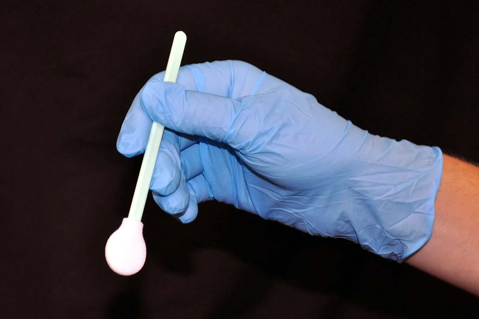 Hand wearing a blue nitrile examination glove holding a swab against a black background